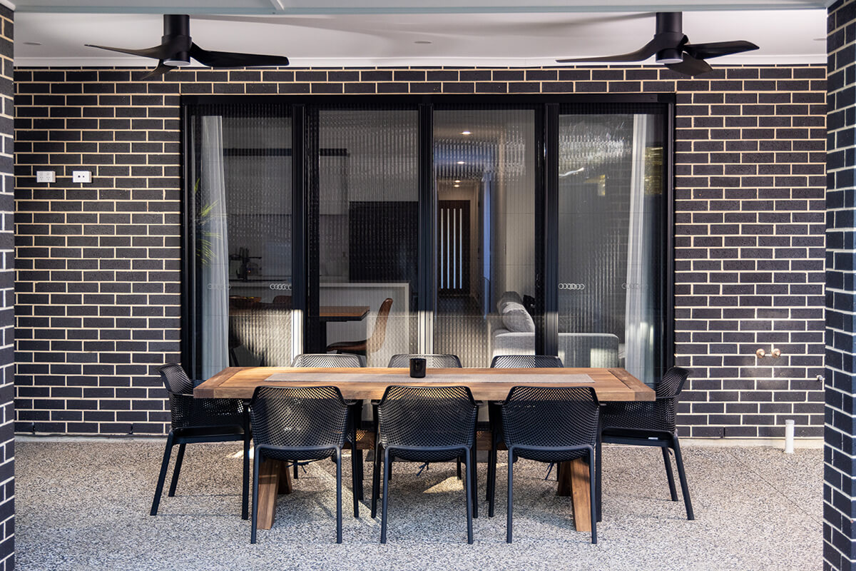Outdoor patio area with dark gray brick walls, a long wooden table with black mesh chairs, and large sliding glass doors to an interior living space.