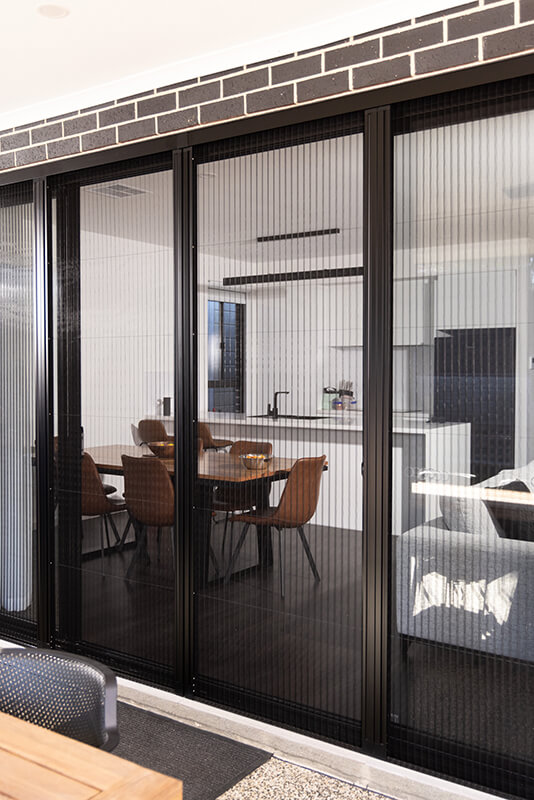 Modern black retractable screen doors on sliding patio doors, separating a dining area with brown chairs from an outdoor patio area.