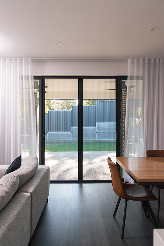 Modern living room with large sliding glass doors to a backyard, showcasing light-colored curtains, a gray sectional sofa, and a light brown wooden dining table with brown leather chairs.