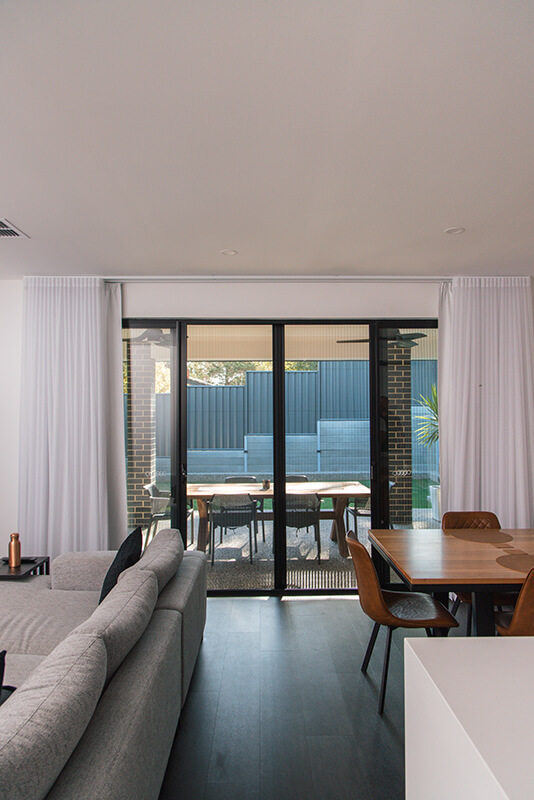 Modern living room with large sliding glass doors to a backyard, showcasing light-colored curtains, a gray sectional sofa, and a light brown wooden dining table with brown leather chairs.