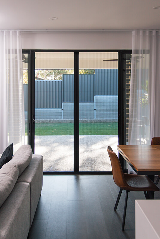 Modern living room with large sliding glass doors to a backyard, showcasing light-colored curtains, a gray sectional sofa, and a light brown wooden dining table with brown leather chairs.