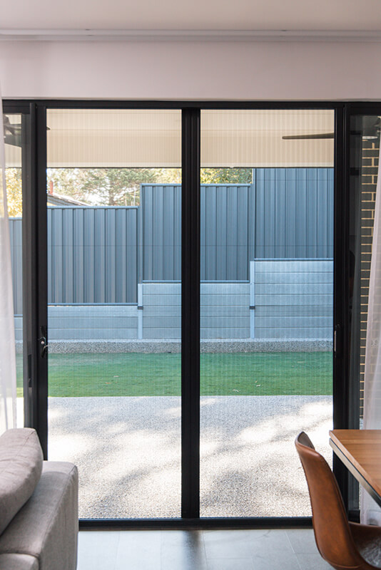 Modern living room with large sliding glass doors to a backyard, showcasing light-colored curtains, a gray sectional sofa, and a light brown wooden dining table with brown leather chairs.