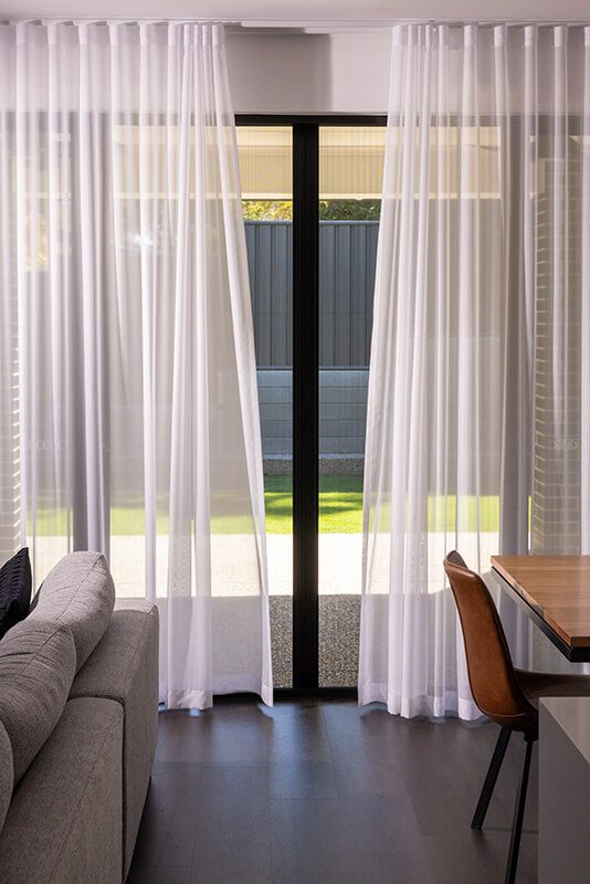 Modern living room with white sheer curtains, a gray sofa, and a view of a backyard through a glass door.