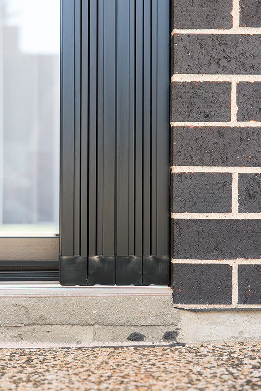 Dark gray sliding door system with vertical slats, mounted against a dark brick exterior wall, with concrete base.
