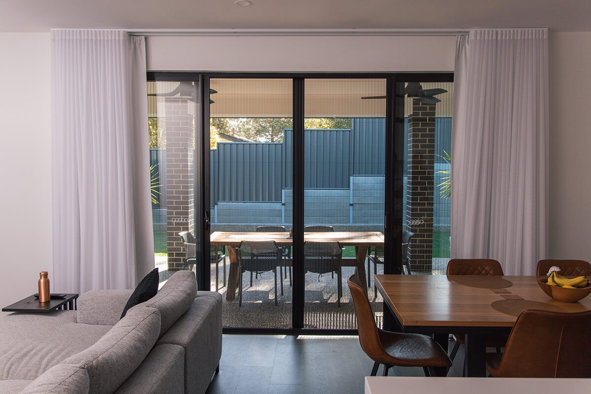 Modern living room with large sliding glass doors to a backyard, showcasing light-colored curtains, a gray sectional sofa, and a light brown wooden dining table with brown leather chairs.