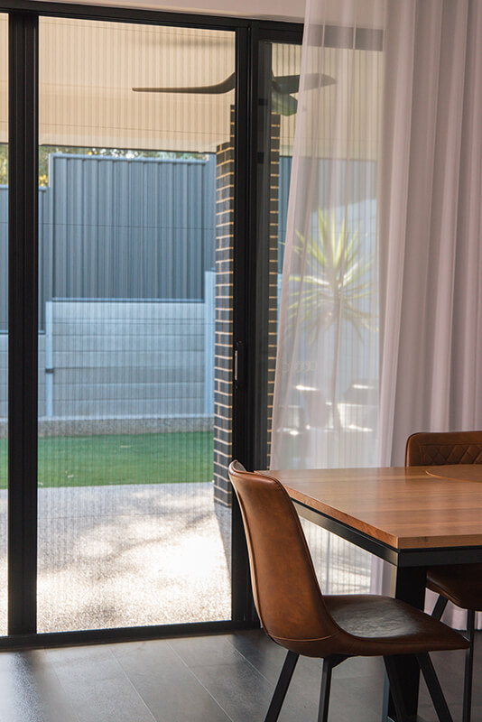 Modern living room with large sliding glass doors to a backyard, showcasing light-colored curtains and a light brown wooden dining table with brown leather chairs.